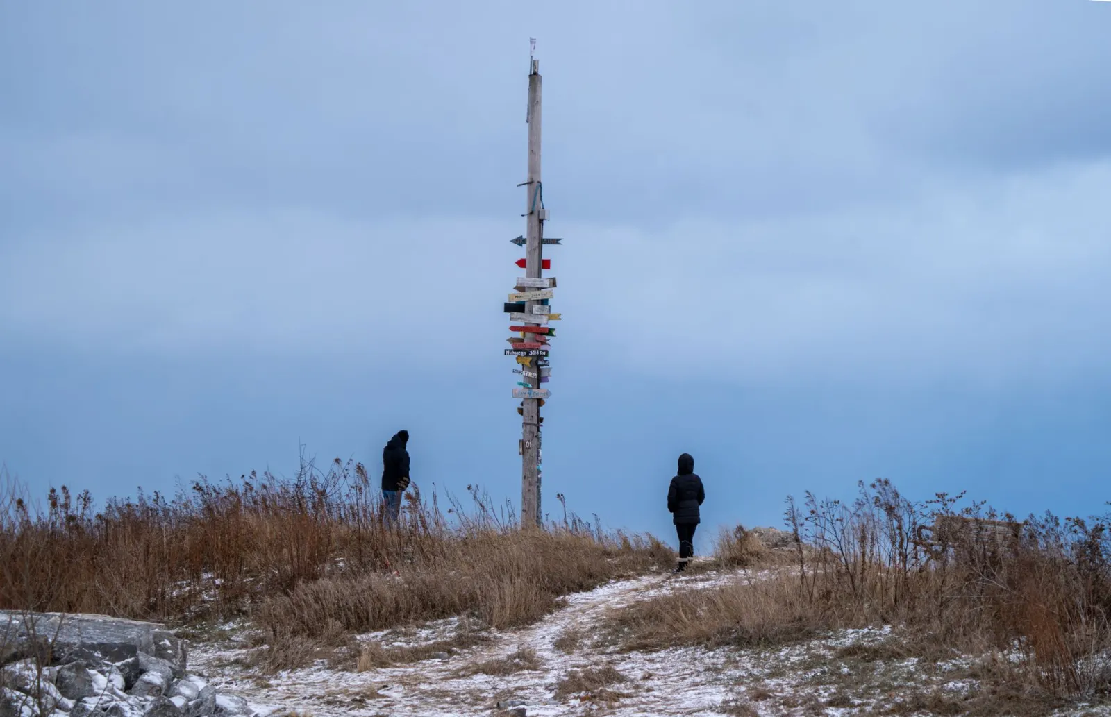 Two people in winter clothing explore a snowy landscape with a directional signpost under a cloudy sky.